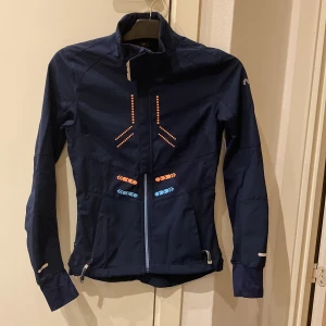 Mörkblå jacka från North Bend - Säljer en mörkblå jacka från North Bend med dragkedja och reflekterande detaljer i orange och blått. Jackan har en hög krage och långa ärmar med skön lång mudd. Fickor på sidorna med dragkedja samt en mindre ficka med dragkedja baktill. Perfekt för utomhusaktiviteter.