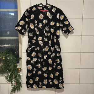 Svart blommig klänning från Camilla Thulin - Säljer en svart klänning från Camilla Thulin med vackert blommönster i vitt och gult. Klänningen har knappar framtill och korta ärmar. Perfekt för en somrig look!