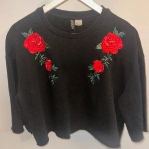 Svart tröja med rosbroderi från H&M - Snygg svart tröja från H&M med vackra röda rosbroderier på bröstet. Tröjan har en avslappnad passform och långa ärmar, perfekt för en trendig look. Passar utmärkt till både jeans och kjol.
