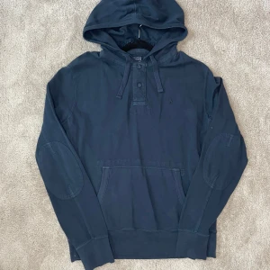 Blå hoodie från Polo Ralph Lauren - Säljer en snygg blå hoodie från Polo Ralph Lauren i storlek M. Den har en klassisk design med knappar framtill och en praktisk magficka. Perfekt för en avslappnad stil och passar bra till både höst och vår. Huvan ger extra mysfaktor! Skicket på hoodie är bra men har ett litet hål på luvan(se sista bilden), skicka gärna prisförslag 
