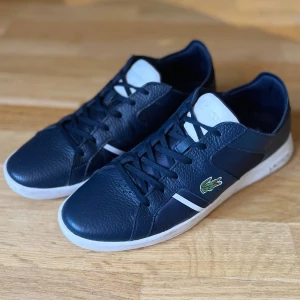 Blå skinnsneakers från Lacoste - Snygga mörkblå sneakers från Lacoste med vita detaljer och klassisk krokodillogga på sidan. Skorna har snörning, en bekväm vit sula och är i bra skick. Perfekta för en stilren look.