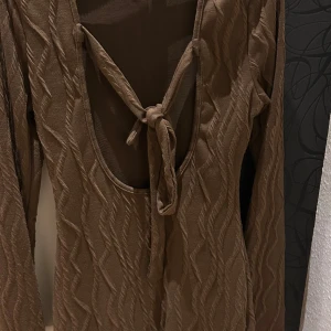 Brun klänning med knytning - Snygg brun klänning med långärmad design och knytning och öppen rygg. Den har ett subtilt mönster som ger en extra touch. Perfekt för en stilren look.