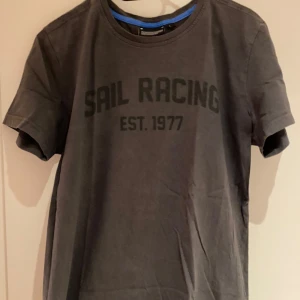 Grå t-shirt från Sail Racing - Snygg grå t-shirt från Sail Racing med tryck 'SAIL RACING EST. 1977' och en storleken L men passar perfekt för M på framsidan. T-shirten har en rund halsringning och ett märke på ärmen. Perfekt för sommar och för en avslappnad stil.