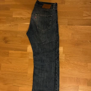 Levis 501 jeans - Klassiska Levis 501 jeans i blå denim med en rak passform. De har en brun läderpatch på baksidan och en ikonisk röd etikett på bakfickan. Perfekta för en tidlös stil.