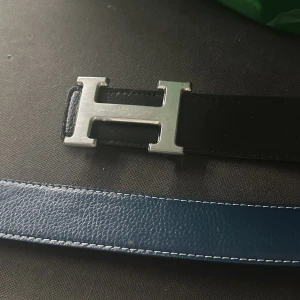 Svart och blått bälte med H-spänne - Snyggt vändbart bälte i svart och blått med ett stilrent H-format spänne i silver. Perfekt för att ge din outfit en lyxig touch. Bältet är gjort av skinn och har en längd på 110 cm.