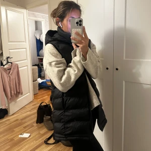 Svart dunväst med huva - Snygg svart dunväst med huva, perfekt för kyliga dagar. Västen har en dragkedja framtill och är quiltad för extra värme. En stilren och praktisk väst för alla tillfällen.