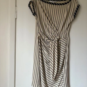 Randig sidenklänning från Diane von Furstenberg - Elegant randig klänning i beige och svart från Diane von Furstenberg. Tillverkad i 100% siden med en knytning i midjan för en smickrande passform. Perfekt för en stilren look.