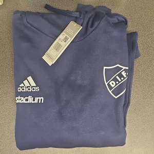 Mörkblå hoodie från Adidas med DIF-logga - Säljer en snygg mörkblå hoodie från Adidas med DIF-logga på bröstet. Tröjan har en klassisk känguruficka och justerbar huva med dragsko. Perfekt för alla DIF-fans! 💙