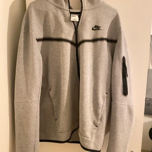 Grå huvtröja från Nike - Säljer en grå huvtröja från Nike med dragkedja och svart logga på bröstet. Tröjan har en praktisk ficka med dragkedja på ärmen och svarta detaljer längs dragkedjan och ärmsluten. Perfekt för en sportig look!