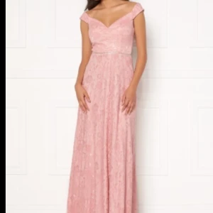 Rosa spetsklänning från Moments New York - Elegant rosa spetsklänning från Moments New York med v-ringning och breda axelband. Klänningen har en glittrig detalj i midjan och en dragkedja i ryggen. Perfekt för speciella tillfällen! Sparsamy använd och inga skador! Klänningen når ner ända till golvet.
