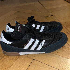 Adidas Mundial Goal - Säljer ett par klassiska Adidas Mundial Goal i svart och vitt. Skorna har en stilren design med de ikoniska tre ränderna på sidan och en gummisula för bra grepp. Perfekta för inomhusfotboll eller casual användning.
