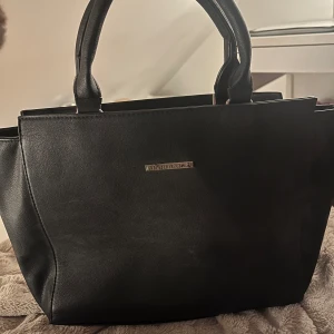 handväska - Elegant svart handväska. Väskan har två robusta handtag och en stilren design med en diskret logotyp på framsidan. Perfekt för att bära dina dagliga nödvändigheter med stil. Extra band tillkommer💓skriv för frågor eller liknande