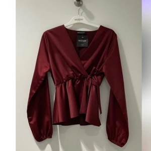 Vinröd omlottblus från Missay - Elegant vinröd omlottblus i satin från Missay. Blusen har långa ärmar och ett knytband i midjan för en smickrande passform. Perfekt för en stilren look eller fest! Toppen är endast använd en gång annars är den helt ny! Fick hem den för två veckor sedan men är tyvärr försent att returnera. Nypris är 299kr