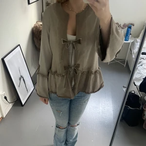 Beige jacka med knyt från by o la la! - Säljer en beige jacka från by o la la med knytband framtill och volangdetaljer. Jackan har långa ärmar och en dekorativ logga på ärmen. Perfekt för en stilren och avslappnad look.