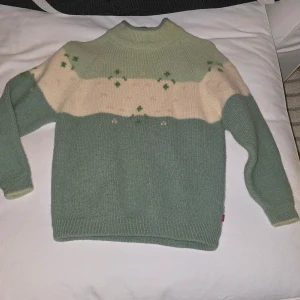 Grön och beige stickad tröja - Mysig stickad tröja i grönt och beige med ett subtilt blommönster. Tröjan har en hög krage och långa ärmar, perfekt för kyliga dagar. Den har en avslappnad passform och ribbade detaljer vid ärmslut och nederkant.