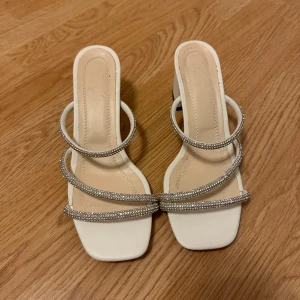 Vita sandaler med glittriga remmar - Eleganta vita sandaletter med fyrkantig tå och glittriga remmar. De har en bred klack som ger stabilitet och stil. Perfekta för en festlig kväll eller en somrig dag.