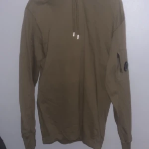 Oliv grön  hoodi från C.P. Company - Säljer en oliv grön hoodi från C.P. Company med huva och långa ärmar. Hoodi har en praktisk ficka på ärmen med en rund detalj. Perfekt för en stilren och avslappnad look. Helt ny plast e kvar