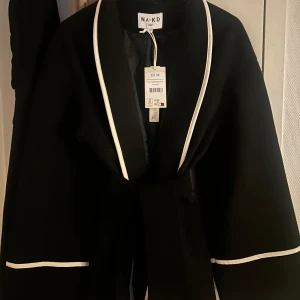 Svart kappa med kontrastband från NA-KD - Ny Snygg svart kappa från NA-KD med vita kontrastband. Den har en knytning i midjan och är perfekt för en enkel outfit, men passar lika bra som till uppklätt. Kappan är långärmad och har en modern design som passar till många tillfällen.  Mjuk och bekväm 