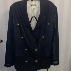 Vintage  kavaj från XIT - Snygg vintage mörkblå kavaj från XIT i storlek 38-40, tillverkad i 100% linne, armar av chiffong.Kavajen har metall guldknappar samt metall spänne på baksidan  och en klassisk design med långärmade ärmar. Perfekt för en stilren look.