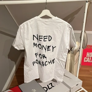 Vit t-shirt med tryck - Säljer en vit t-shirt med texten 'Need Money for Porsche' tryckt på baksidan. T-shirten har en klassisk rund hals och korta ärmar. Perfekt för en avslappnad stil med en humoristisk touch.