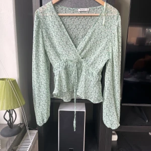 Grön blommig omlottblus från Stradivarius - Säljer en söt grön blommig omlottblus från Stradivarius. Blusen har långa ärmar och ett knytband i midjan för en fin passform. Ett hål och skadad vid axeln
