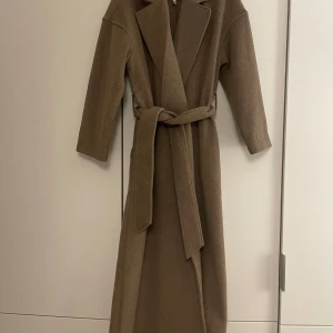 Beige kappa med bälte - Världens snyggaste kappa. Den är lång (maxi) och går att styla med allt. Har två fickor och band vid midjan. Har använt den Max 3 gånger. Säljer då den ej kommer till användning. Nypris är 1600 kr inköp på Ellos. Jag är 165 och på bilderna ser ni hur kappan är på mig med klackar på. 40% Ull 40% polyester och 20% viskos