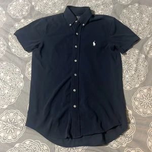 Ralph Lauren pike  - Säljer en stilren mörkblå kortärmad skjorta från Ralph Lauren. Skjortan har en klassisk button-down krage och är dekorerad med det ikoniska broderade logotypen på bröstet. Perfekt för en avslappnad men ändå elegant look.