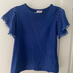 Blå topp med pärldetaljer från Savida - Snygg blå topp från Savida med korta ärmar och vackra pärldetaljer. Ärmarna har ett unikt mönster med spets och pärlor som ger en elegant touch. Perfekt för att lyfta vilken outfit som helst.