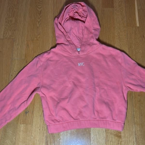 Rosa hoodie från H&M med NYC-tryck - Säljer en snygg rosa hoodie från H&M med ett stilrent 'NYC' tryck på bröstet. Perfekt för en avslappnad look. Hoodien har långa ärmar och en bekväm huva.