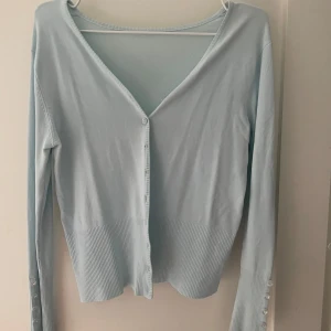 Ljusblå kofta med knappar - Snygg ljusblå kofta med ribbade detaljer och knappar framtill. Perfekt för lager-på-lager och ger en stilren look. Långärmad design med en v-ringad halslinje.