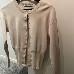 Beige kofta från Zara - Säljer en stilren beige kofta från Zara med knappar framtill. Koftan har långa ärmar och ribbade kanter vid ärmslut och nederkant. Oanvänd 