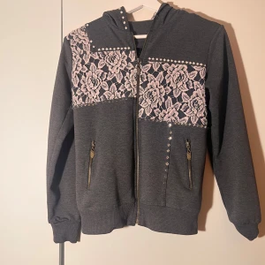 Grå kofta med spetsdetaljer - Snygg grå hoodie med dragkedja och spetsdetaljer på bröstet och huvan. Tröjan har långa ärmar och nitar vid axlarna och fickorna. Säljer då den är för liten på mig, annars hade den sett jättesnygg ut med ett par lågmidjade jeans. Saknar några stenar därav priset