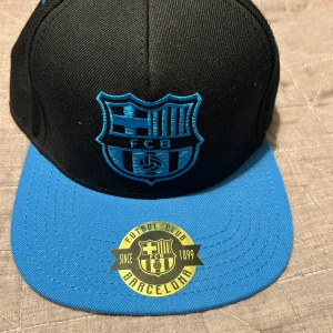 Svart och blå keps från FC Barcelona - Snygg svart och blå keps med FC Barcelona-logga framtill. Kepsen har en platt skärm och är tillverkad i en blandning av akryl och ull. Perfekt för fotbollsfans som vill visa sitt stöd för laget.   Jag köpte denna keps i barcelona 2016-17 inne i camp nou och har använt den kanske 1-2 gånger och som ni se rop bilden är den i toppskick 