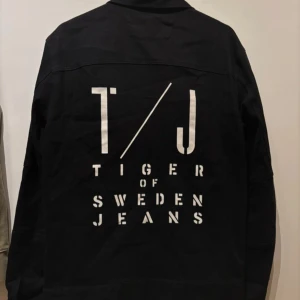 Svart jeansjacka från Tiger of Sweden - Snygg svart jeansjacka från Tiger of Sweden med tryck på ryggen. Jackan har knappar framtill och två bröstfickor. Perfekt för en stilren look. Oanvänd i storlek M. 