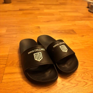 Svarta sandaler från Maya Delorez - Snygga svarta sandaler från Maya Delorez med logotyp på ovansidan. Perfekta för en avslappnad stil och bekväma att bära. Sandalerna har en enkel design med en bred rem över foten. Orginal pris 350