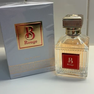 B Rouge Eau de Parfum - Elegant parfymflaska med en lyxig design. Flaskan är genomskinlig med ett rött och guldigt emblem. Förpackningen har en stilren vit och guldig design med detaljer av en kristallkrona och pelare. Perfekt för den som söker en sofistikerad doftupplevelse.