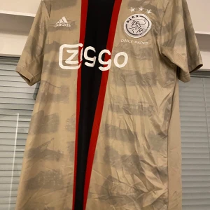 Ajax fotbollströja från Adidas - Snygg beige fotbollströja från Adidas med Ajax-logga och sponsortryck. Tröjan har korta ärmar och en unik design med röda och svarta detaljer längs framsidan. Perfekt för fotbollsfans som vill sticka ut.