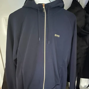 Mörkblå hoodie från hugo boss - Snygg mörkblå hoodie från Boss med dragkedja och justerbar huva. Perfekt för en avslappnad stil. Den har en diskret logga på bröstet och långa ärmar.storlek L