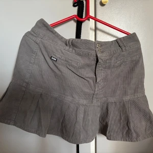 Grå manchesterkjol från Minga London - Snygg grå manchesterkjol från Minga London med volangdetalj. Kjolen har en kort längd och stängs med knappar framtill. Perfekt för en trendig look!