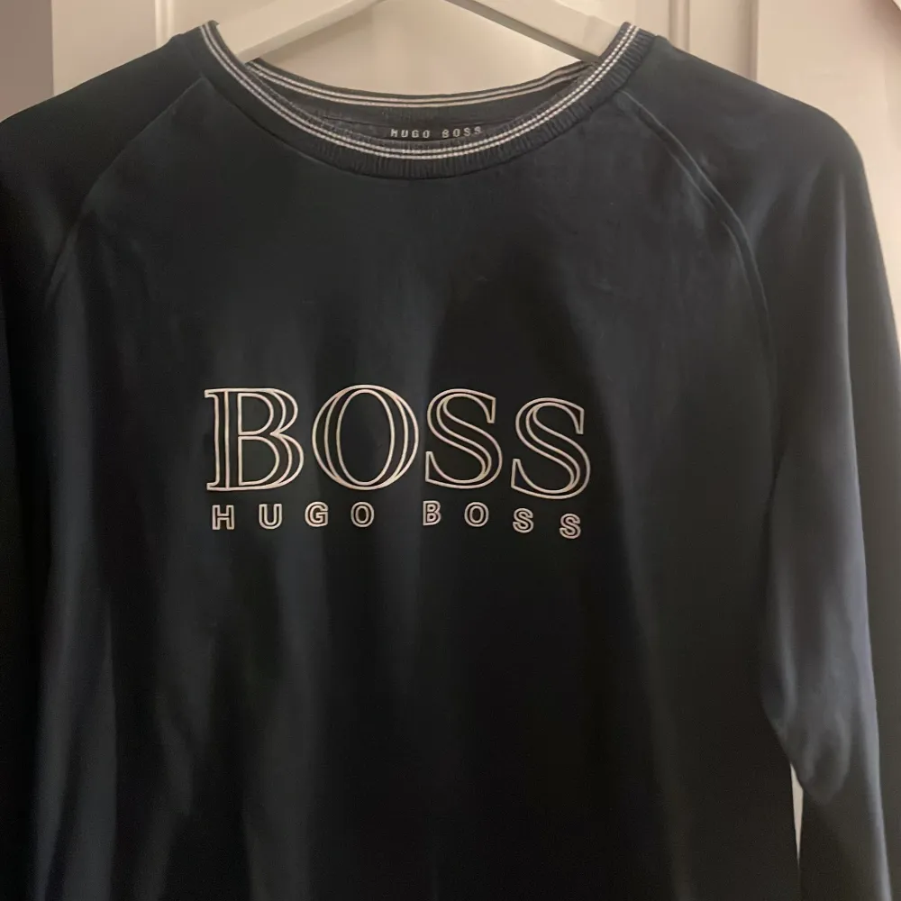 Snygg svart tröja från Hugo Boss med stort BOSS-tryck på framsidan. Tröjan har långa ärmar och en rund halsringning med diskreta ränder. Perfekt för en stilren look.. Neuletakit & Villapaidat.