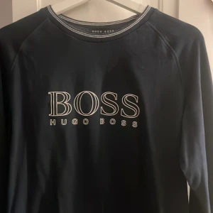 Svart tröja från Hugo Boss - Snygg svart tröja från Hugo Boss med stort BOSS-tryck på framsidan. Tröjan har långa ärmar och en rund halsringning med diskreta ränder. Perfekt för en stilren look.