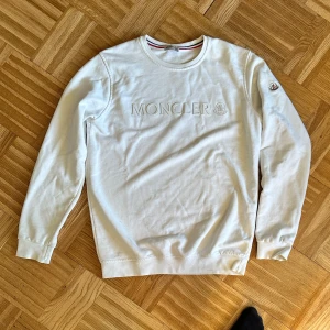 Vit sweatshirt från Moncler - Säljer en stilren vit sweatshirt från Moncler. Den har ett broderat Moncler-logotyp på bröstet och en liten logga på ärmen. Tröjan är långärmad och perfekt för både vardag och lite finare tillfällen. Superbekväm och i bra skick! Storlek S-M