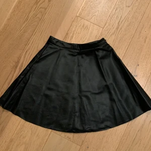 Svart kjol från NA-KD - Snygg svart kjol i väldigt mjuk skinnimmitation från NA-KD i storlek 34. Kjolen har en stilren design och är perfekt för både vardag och fest. Den har en dragkedja i sidan för enkel på- och avtagning.