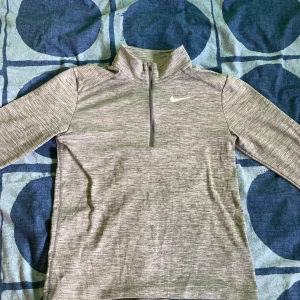 Grå långärmad Nike sportsfleece  - Säljer en grå långärmad sport tröja från Nike med halv dragkedja. Perfekt för träning eller en avslappnad dag. Tröjan är i bra skick och har en stilren design med Nike-loggan på bröstet. Jätteskön och i perfekt skick! 