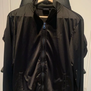 Svart jacka från Emporio Armani - Säljer en stilren svart jacka från Emporio Armani. Den har en dragkedja framtill och är perfekt för både vardag och lite finare tillfällen. Jackan är i bra skick och har en snygg, enkel design med EA7-loggan på bröstet.