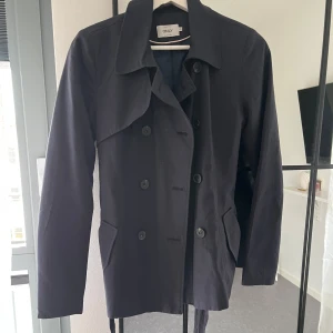 Svart kappa från ONLY - Säljer en stilren svart kappa från ONLY. Den har en klassisk design med knappar framtill och ett bälte i midjan för en snygg passform. Perfekt för höst och vår. Jackan är i bra skick och är knappt använd!