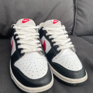 Nike Dunk i svart, vitt och rött -  snygga Nike Dunk sneakers i svart, vitt och rött. Skorna har en klassisk design med vit bas och svarta överlägg, samt en röd Nike-logga som verkligen poppar. Perfekta för både vardag och fest! De är i bra skick bara ett pyttelitet hål i hälen