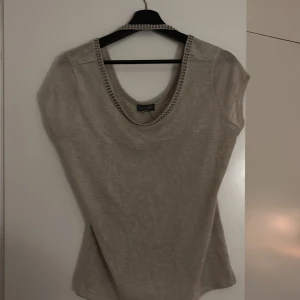 Beige topp från Essentials Milano - Säljer en stilren beige topp från Essentials Milano. Toppen har en rund halsringning med dekorativa detaljer och är ärmlös. Perfekt för en avslappnad look.