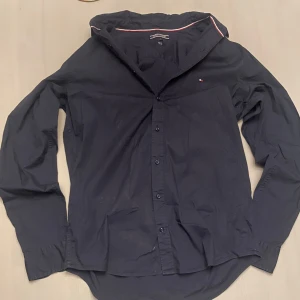 Svart skjorta från Tommy Hilfiger - Snygg svart skjorta från Tommy Hilfiger i slim fit med stretch. Skjortan har långa ärmar och knappar framtill. Perfekt för en stilren look. Inget pris är spikat i sten!