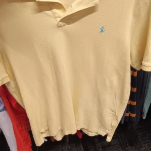 Gul pikétröja från Ralph Lauren - Säljer en klassisk gul pikétröja från Ralph Lauren med blå logga på bröstet. Tröjan har en klassisk passform och är perfekt för en avslappnad stil. Den är tillverkad i ett bekvämt material som passar bra till både vardag och fritid.